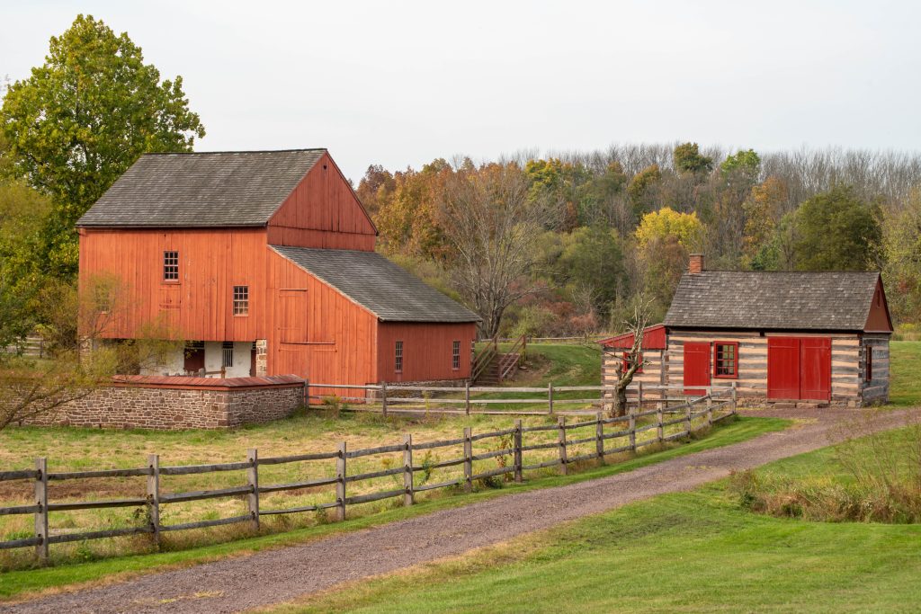 Historic colonial American farm scene, dirt road, log cabin and red barn on the historic Daniel Boone Homestead in Pennsylvania, autumn landscape with yellow and orange leaves and copy space.